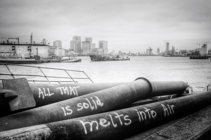 "All that is solid melts into air" graffitti on old pipes lying alongside the River Thames, London. Black and white photo by Su Leslie, 2015.
